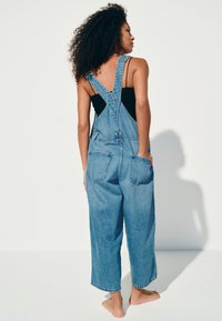 Femeie cu păr creț stă desculță cu fața spre spate, purtând o salopetă albastră din denim și un top negru fără mâneci pe un fundal alb simplu.