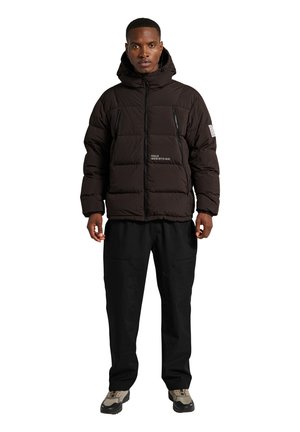 Mand iført sort dunjakke med hætte, sorte bukser og beige vandresko, stående med front mod kamera på hvid baggrund.