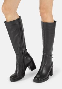 Bottes noires montant jusqu'aux genoux en cuir lisse, avec un talon épais, un bout arrondi, une fermeture éclair latérale et un discret logo sur le talon.