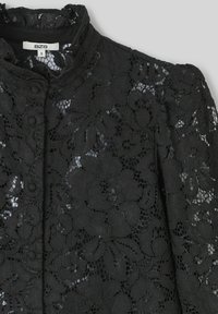 Tissu en dentelle noire avec motif floral, col montant et fermeture à boutons. Surface texturée avec des éléments transparents contrastants.