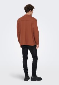 Άνδρας με σγουρά μαλλιά, γυρισμένος πίσω, φοράει μακρυμάνικη μπλούζα σε σκουρόχρωμη σκουριά, μαύρο τζιν και μαύρα μποτάκια, σε απλό φωτεινό φόντο.