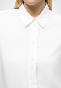 Fehér, gombos ing klasszikus gallérral. Simaságot biztosító anyagból készült, egységes gombos, elől elvarrt gallérúval és laza szabással.