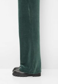 Grüne Cord hosen mit weitem Bein und gerippter Textur, kombiniert mit schwarzen, klobigen Stiefeln. Fokus auf Stoff und Form.