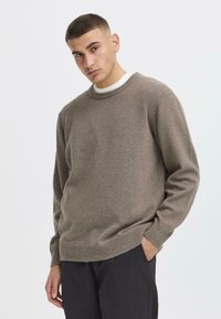 Brown knit sweater with a round neckline and ribbed cuffs, featuring a soft texture and a relaxed fit. Worn over a white shirt.