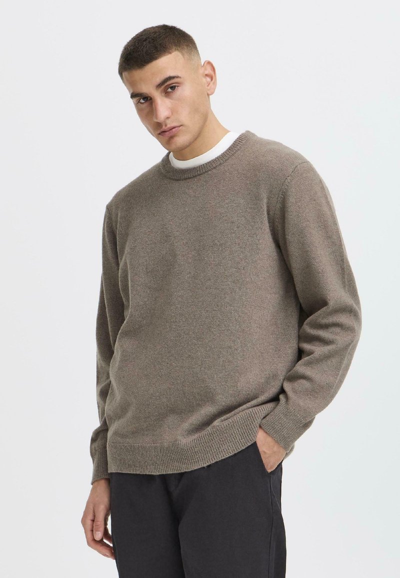 Brown knit sweater with a round neckline and ribbed cuffs, featuring a soft texture and a relaxed fit. Worn over a white shirt.