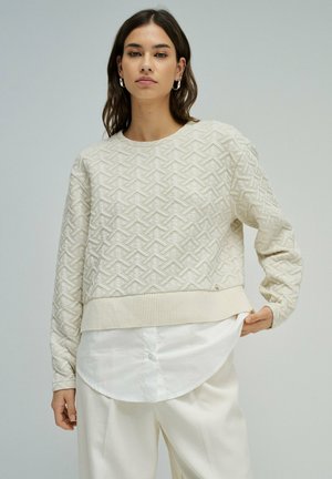 Mujer con un suéter crema de textura, de manga larga, sobre una camisa blanca abotonada y pantalones blancos, de pie contra un fondo liso.