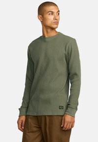 Olivegrünes langärmeliges Thermoshirt mit strukturiertem Rippmuster, Rundhalsausschnitt und Logopatch unten links. Getragen mit braunen Hosen.