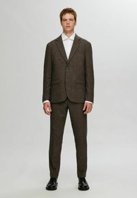 Barna tweed öltöny, strukturált zakóval, bemetszett gallérral, egygombos záródással és egyenes lábú nadrággal. Fehér galléros inggel párosítva.