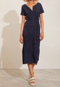 Donna che indossa un vestito a maglia blu navy con bottoni e maniche corte, con una cintura in vita, abbinato a sandali bianchi con platform, in piedi su un pavimento neutro.