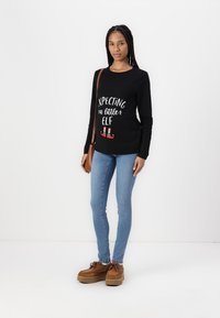 Black long-sleeve shirt with white text reading “Expecting a little elf,” paired with blue jeans and brown platform shoes.