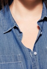 Koszula dżinsowa w jasnoniebieskim kolorze, z zapięciem na guziki z przodu, kołnierzem i dwoma kieszeniami na piersi. Materiał jest miękki, z widocznymi detalami szycia.