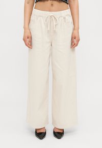Cream wide-leg pants with a drawstring waist, featuring two side pockets and a smooth texture. Paired with black pointed-toe shoes.