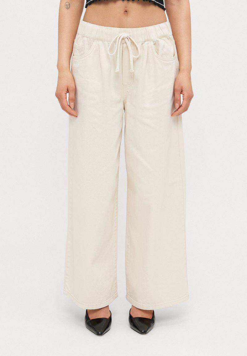 Cream wide-leg pants with a drawstring waist, featuring two side pockets and a smooth texture. Paired with black pointed-toe shoes.