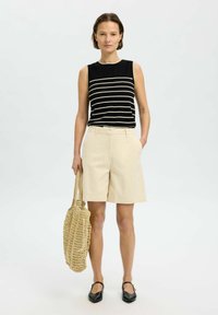 Top senza maniche a righe bianche e nere abbinato a pantaloni beige su misura. Il modello tiene una borsa intrecciata e indossa scarpe nere.