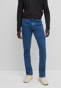 Jeans de denim azul con un corte recto, con un diseño clásico de cinco bolsillos y un lavado medio. Combinados con zapatillas en crema y una camisa negra de manga larga.