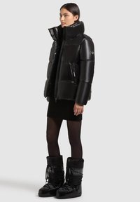 Melnā pūšamā jaka ar augstu apkakli, spīdīgu tekstūru un pūstām sekcijām. Apvienota ar melniem svārkiem un augstiem melniem zābakiem ar auklām.