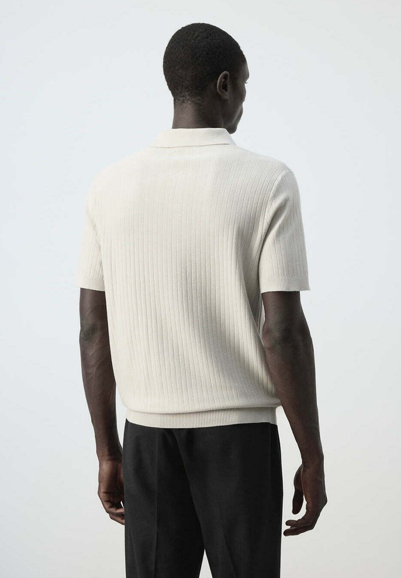 Homme à la peau foncée portant un t-shirt blanc à manches courtes en maille côtelée et un pantalon noir, debout de dos sur un fond clair uni.