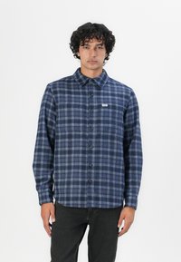 Camisa de franela a cuadros azul marino con botones, con cuello y una pequeña etiqueta de logo en el pecho, combinada con jeans oscuros.