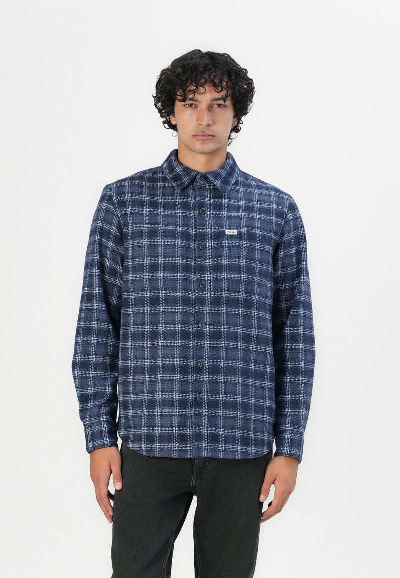 Camisa de franela a cuadros azul marino con botones, con cuello y una pequeña etiqueta de logo en el pecho, combinada con jeans oscuros.