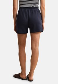 Person trägt marineblaue Shorts mit elastischem Bund, gestreiftes Oberteil und braune, gewebte Slipper, von hinten stehend vor weißem Hintergrund gezeigt.