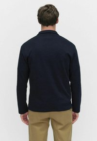 Mörkblå långärmad skjorta med polo-krage och ribbad struktur, sedd bakifrån, bärs med khakifärgade byxor. Inga synliga mönster eller detaljer.