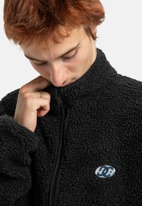 Zwarte fleece jas met een geweven, pluizige buitenkant, hoge kraag en rits aan de voorkant. Bevat een ronde logo patch aan de linkerzijde.