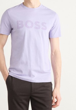 Homme portant un t-shirt violet clair à manches courtes avec le mot "BOSS" imprimé sur la poitrine et un pantalon noir sur un fond uni.