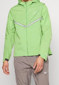 Veste zippée légère verte avec capuche, détails à bandes réfléchissantes et poignets en tricot. Portée avec un pantalon gris orné d'un logo.