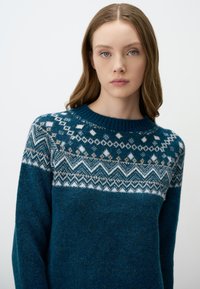 Teal gebreide trui met een ronde halslijn, een geblokte yoke met witte en beige geometrische patronen, en een zachte, geweven afwerking.