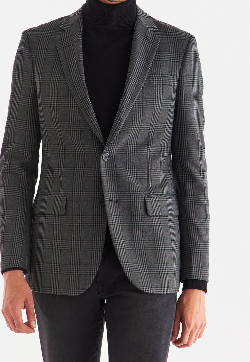 Homme portant un blazer gris à carreaux ajusté par-dessus un col roulé noir et un pantalon noir, debout sur un fond clair uni.