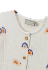 Haut en tissu blanc texturé avec des arc-en-ciel et des escargots brodés colorés, avec des boutons en bois sur le devant. Étiquette de marque visible.