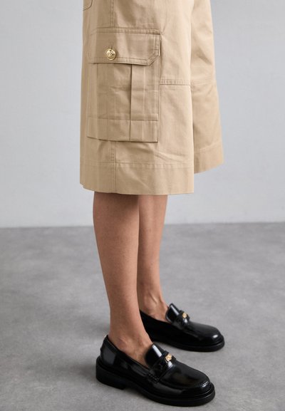 Shorts cargo beiges avec poches latérales et accessoires en or, associés à des mocassins noirs brillants avec un accent décoratif. Texture lisse.