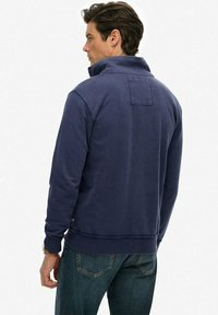 Granatowy sweter z wysokim kołnierzem i naszywką z tyłu. Wykonany z miękkiej tkaniny, z długimi rękawami i ściągaczami przy dolnym brzegu.