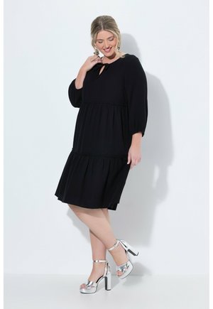 Donna che indossa un vestito nero al ginocchio con maniche 3/4 e sandali argento con tacco alto, in piedi su uno sfondo bianco.