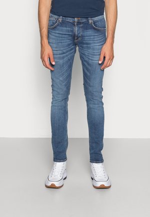 Homme portant un jean bleu slim, des baskets blanches et une chemise bleu foncé, debout devant un fond uni clair.