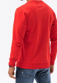 Sudadera roja con cuello redondo, puños y dobladillo acanalados. La tela parece suave y lisa, combinada con jeans azules desgastados.