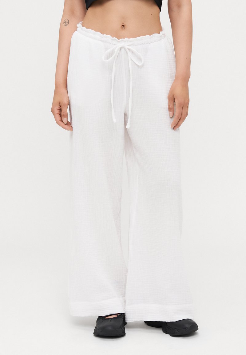 White wide-leg pants with a textured fabric, elastic waistband, and drawstring. Hem features a subtle, solid trim. Paired with black shoes.