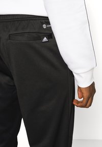 Černé atletické kalhoty s přiléhavým střihem, bočními kapsami a logem Adidas na pase. Vyrobené z odolné tkaniny s hladkým textura.