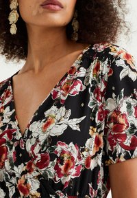 Blommig svart svepklänning med röda och krämfärgade blommor, korta puffärmar och samlad detalj vid halslinjen. Tillbehör: stora örhängen.