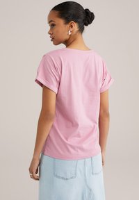 Tricou roz din bumbac cu mânecă scurtă, cu tivuri răsucite și o croială relaxată, asortat cu o fustă din denim deschis. Vedere din spate.
