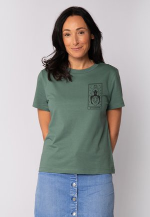 Groene katoenen t-shirt met korte mouwen heeft een zwart grafisch print van een tarotkaart in het midden van de borst, met een leeuw en het woord "KRAACHT."