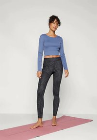 Nő mezítláb áll egy rózsaszín jógaszőnyegen, kék hosszú ujjú crop topot és fekete mintás leggingset visel, sima háttér előtt.