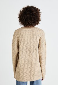Beige strikket genser med et strukturelt kabelmønster; har en rund halslinning og senkede skuldersømmer, sammen med lyseblå jeans.