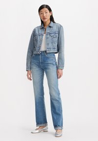 Chaqueta de mezclilla azul claro con dos bolsillos en el pecho, combinada con jeans altos de cintura azul claro y bailarinas plateadas con detalles de lazo.