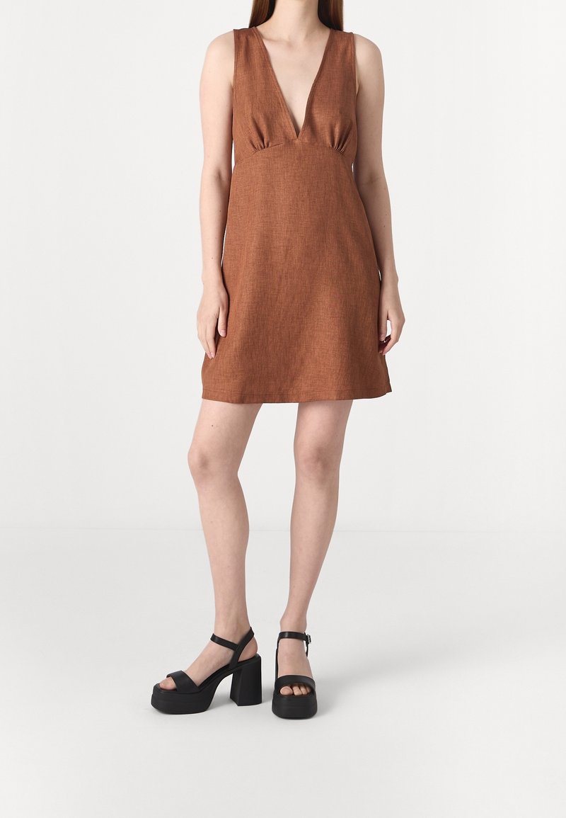 Femme portant une robe sans manches couleur rouille avec un profond décolleté en V et des sandales à plateforme noires épaisses, debout sur un fond uni clair.