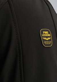 Tissu noir avec un patch jaune et noir portant l'inscription PME Legend American Classic avec un emblème d'aigle.