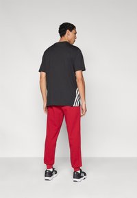 Μαύρο T-shirt με κοντά μανίκια, συνδυασμένο με κόκκινο αθλητικό παντελόνι. Φέρει τρεις λευκές ρίγες κατά μήκος της πλευράς του παντελονιού. Μαύρα αθλητικά παπούτσια.