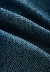 Tissu en denim bleu avec un motif de tissage diagonal, plié en vagues douces créant des effets de lumière et d’ombre texturés.