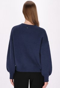 Pulover tricotat bleu marin, cu o croială lejeră, umeri lăsați, manșete și tiv cu nervuri. Textură moale cu un mic detaliu logo pe spate.