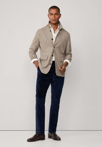 Smeđa jakna od vune s visokim ovratnikom, prednjim gumbima i dva džepa; uparena s tamnoplavim kordura hlačama i smeškim kožnim cipelama.